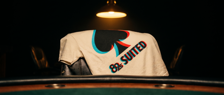A cream 89 Suited tee with a spade motif, laid across the rail of a green poker table