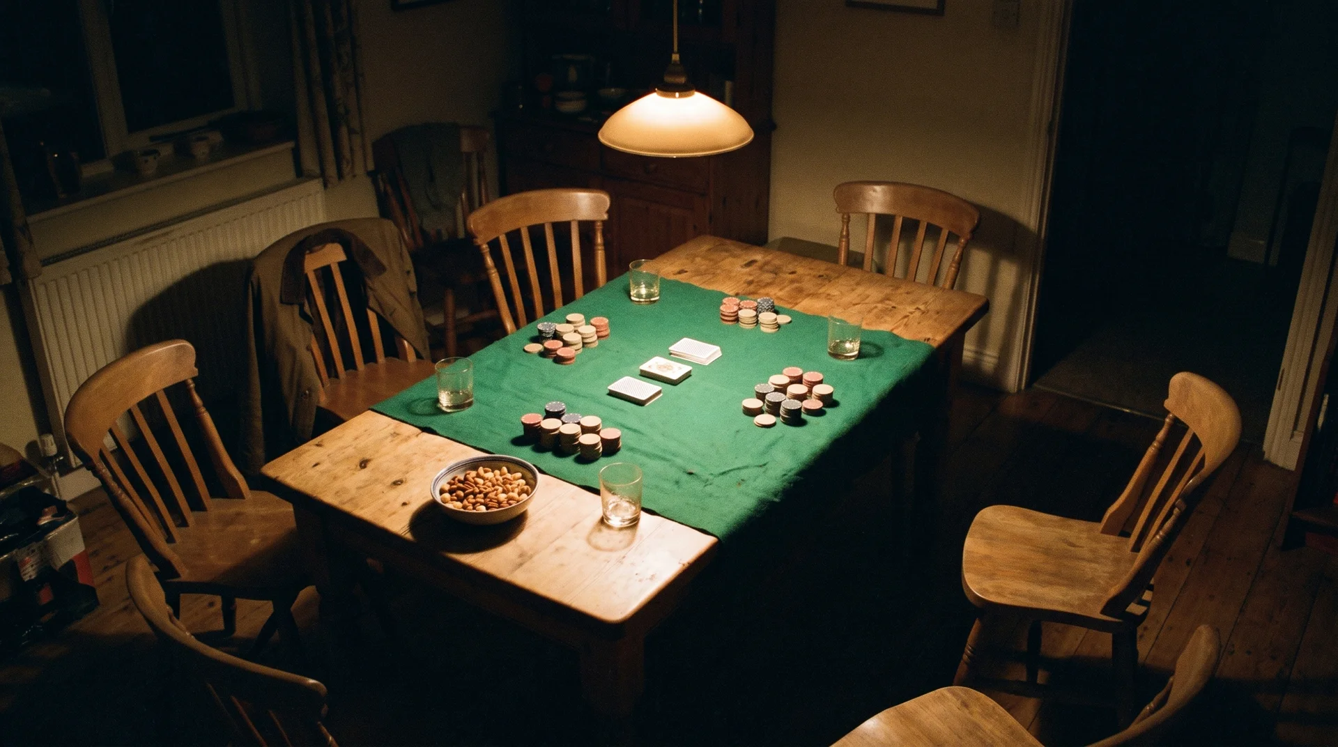 A kitchen table under a single pendant lamp, green felt thrown over the wood, chips in uneven stacks and a bowl of nuts, five empty chairs pulled at angles.