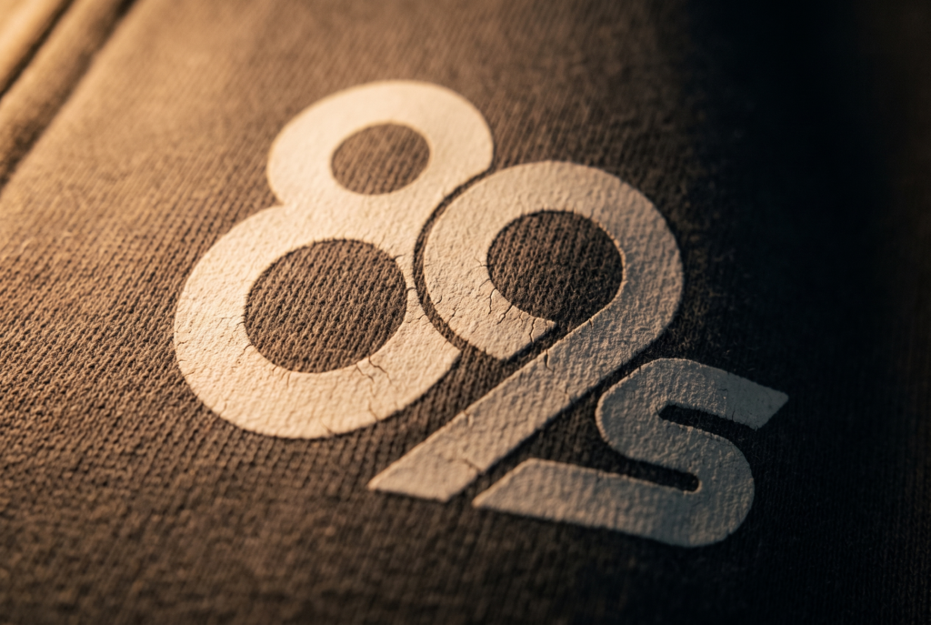 The 89s monogram in cream on dark fabric, lit from the side, close enough to see the weave.