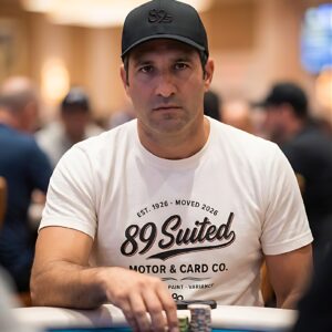A poker player in the cream Motor & Card Co. tee, looking straight at the camera across the felt, a cap with the 89s monogram.
