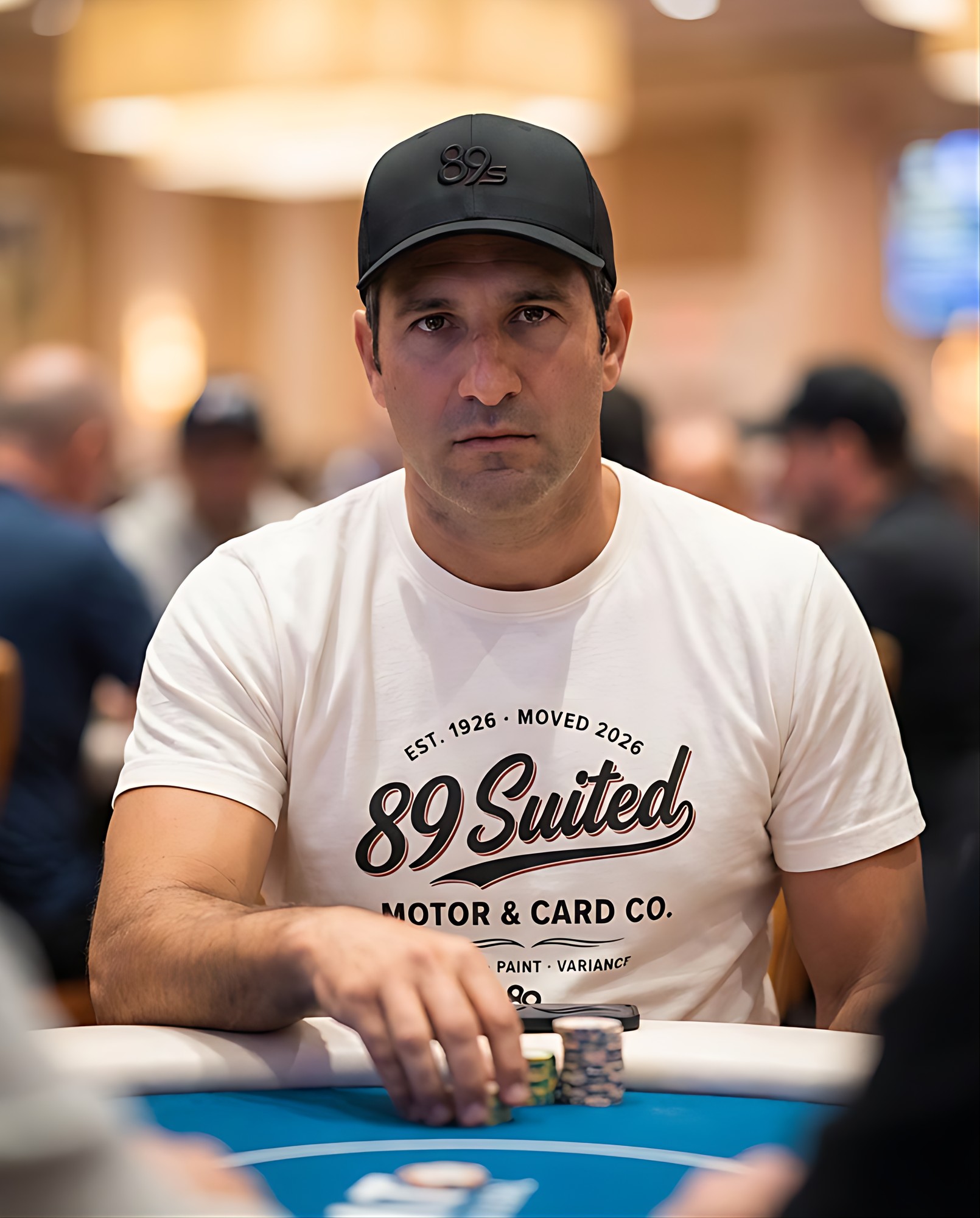 A poker player in the cream Motor & Card Co. tee, looking straight at the camera across the felt, a cap with the 89s monogram.