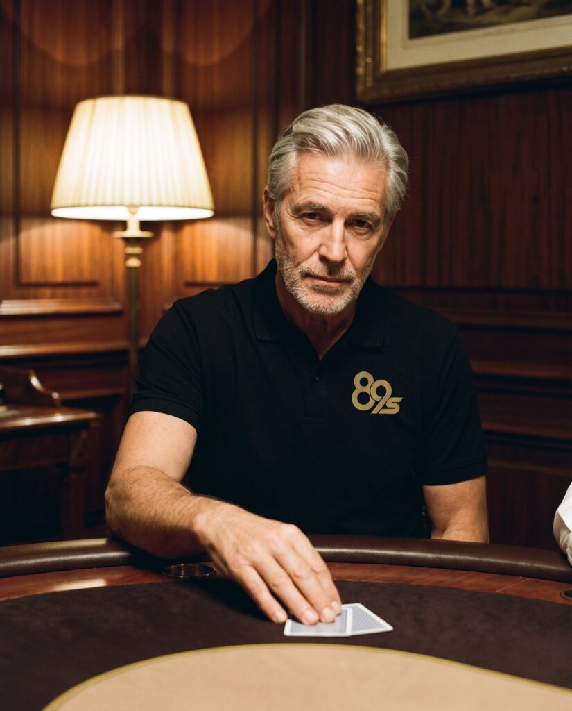 A silver-haired man in the black 89s Polo seated at a poker table in a wood-panelled room, two cards under his hand.